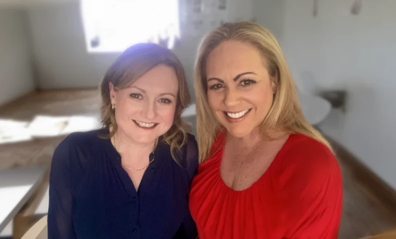 Two women smiling and sitting together in a training room. Rebecca from Targeted Training is on the left, wearing a navy blue blouse, and Christine from Targeted Training is on the right, wearing a red blouse. The background is blurred, showing a bright window and light-coloured furniture.